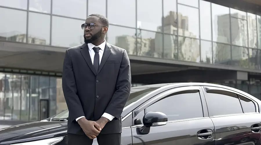 a man wears a suit and stands with car -condor security Executive Protection Toronto (Security Services)